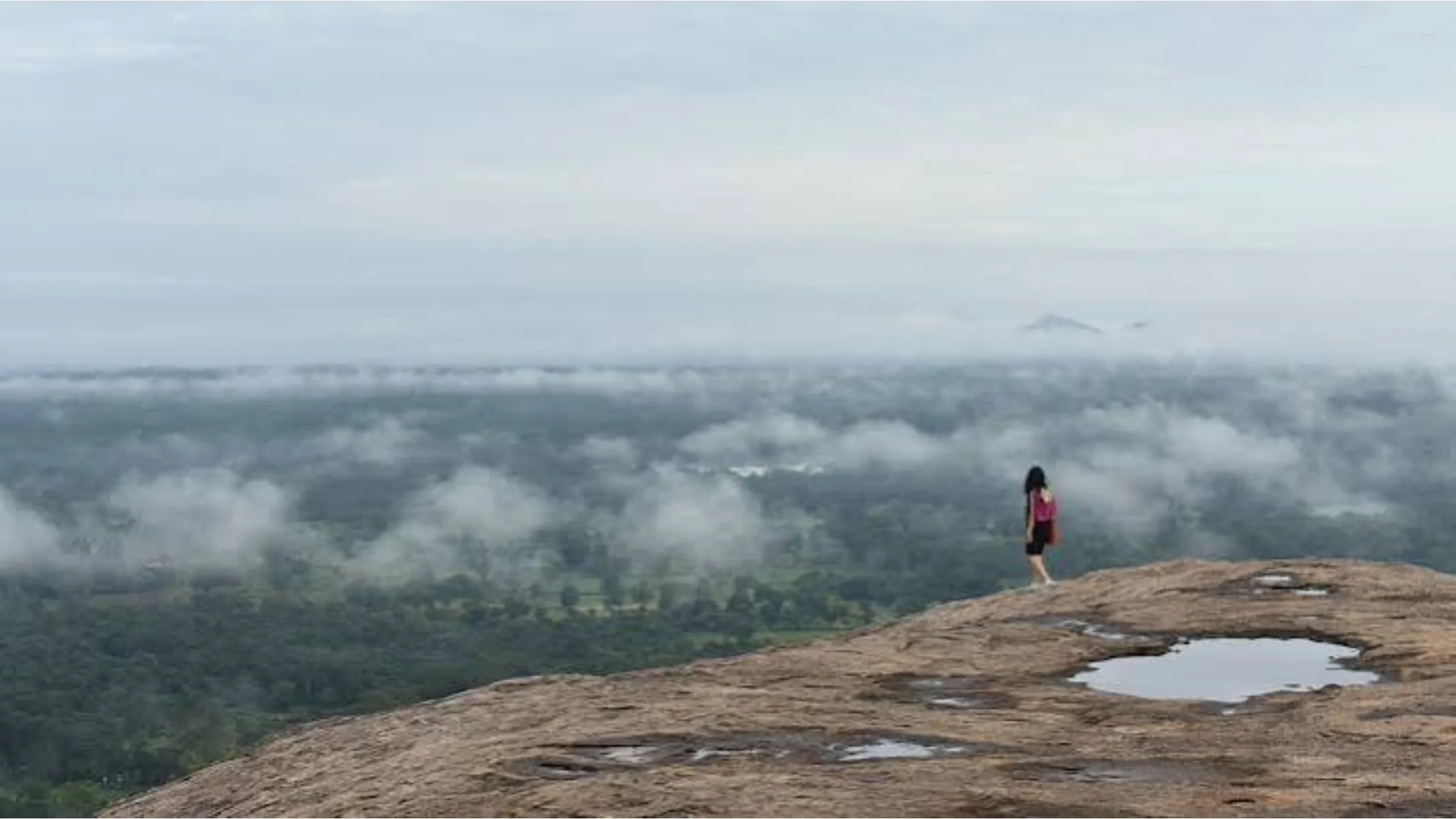 A hiker standing at the edge of a misty highland plateau overlooking forest below