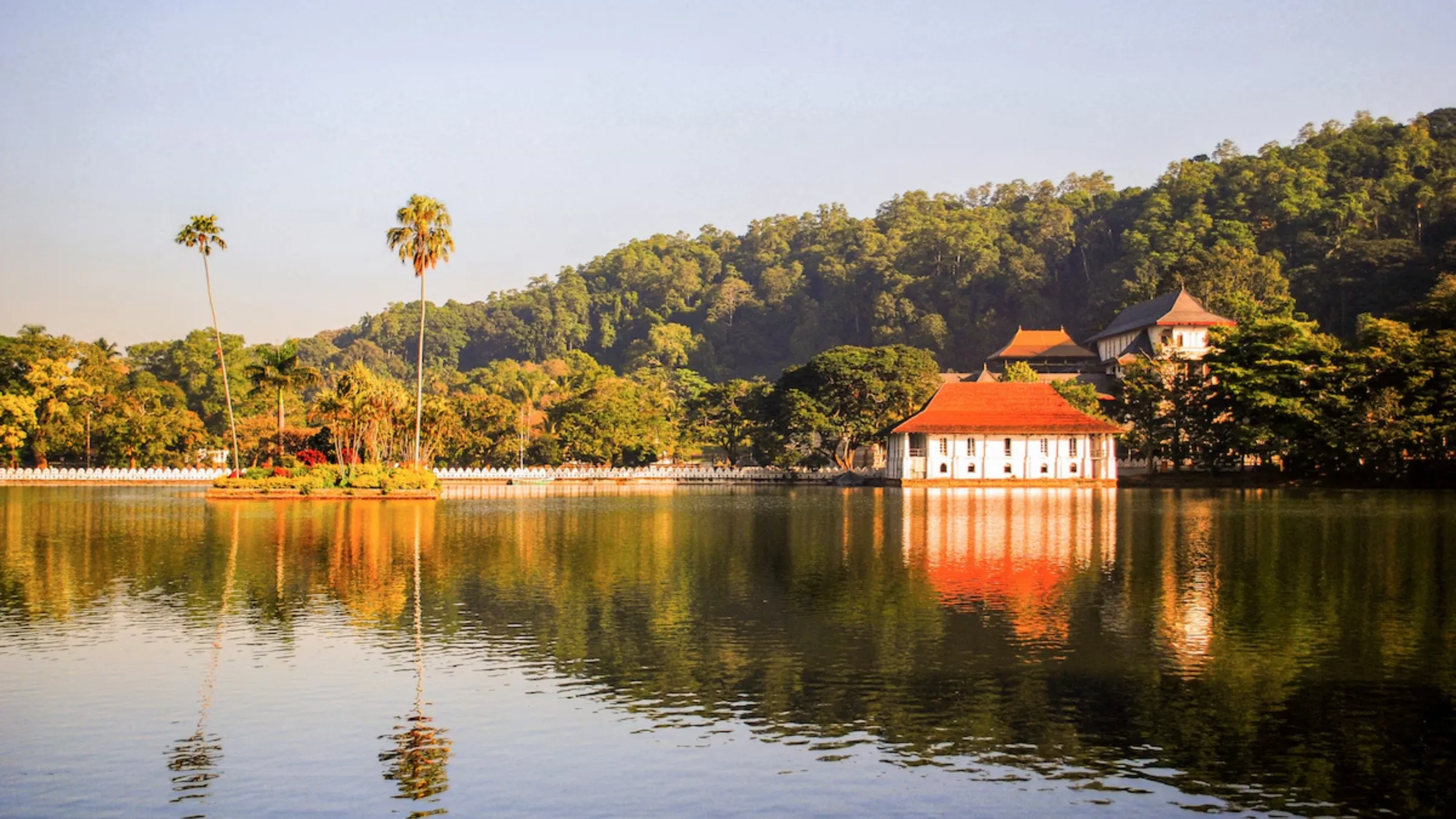 The Royal Bath's white and orange-roofed pavilion reflected in Kandy Lake at golden hour, framed by palm trees